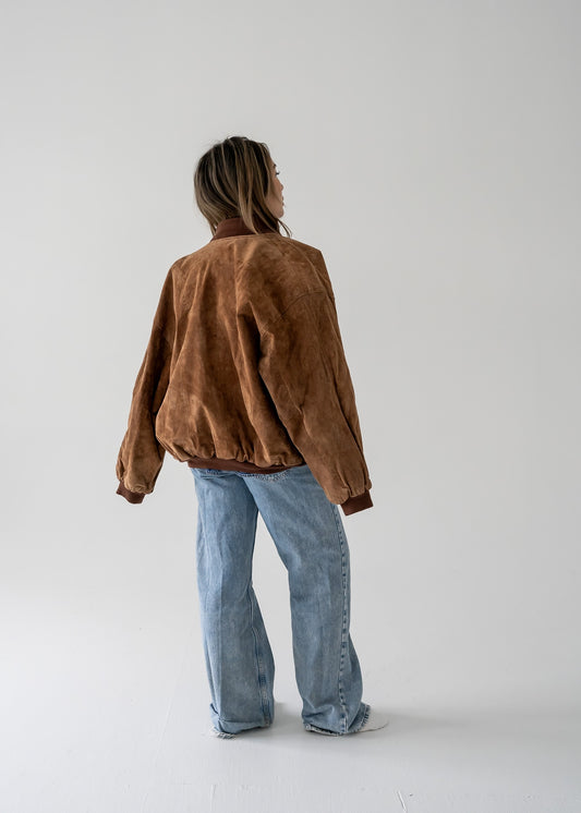 A person wearing a vintage brown suede leather bomber jacket with a mandarin collar, over a blue denim shirt and blue jeans