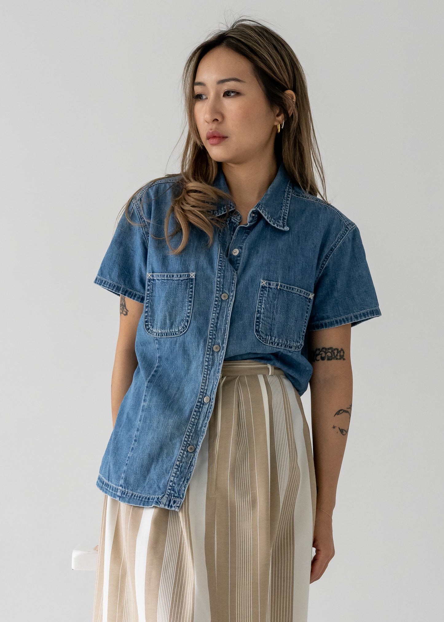 Person wearing a vintage medium wash denim short sleeve shirt and beige and white skirt against a plain background