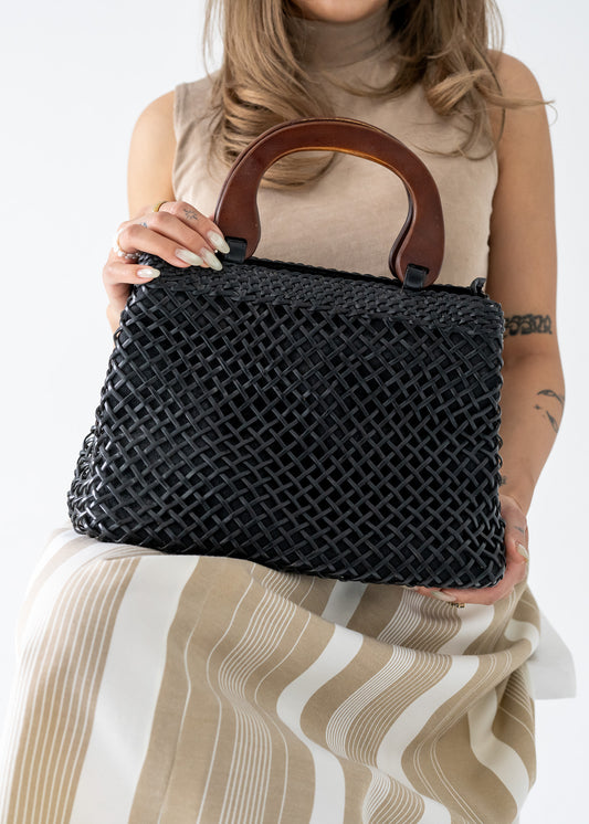 Person holding a vintage black leather handbag with an open basket weave pattern and wooden handles against a neutral background