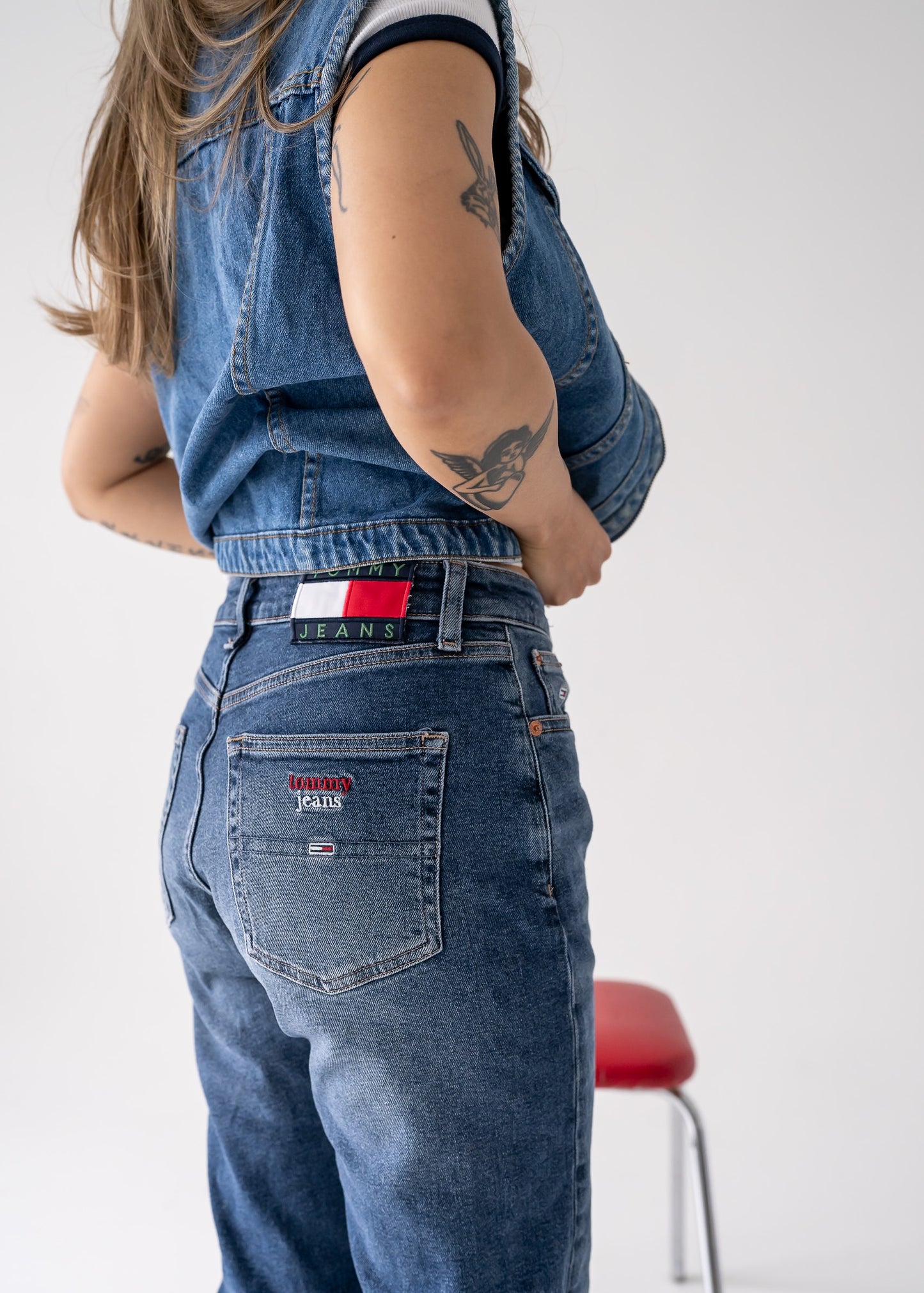 A person wearing a vintage collared zip front medium wash denim vest with buttoned flap pockets on the breast and blue jeans over a white tank top.