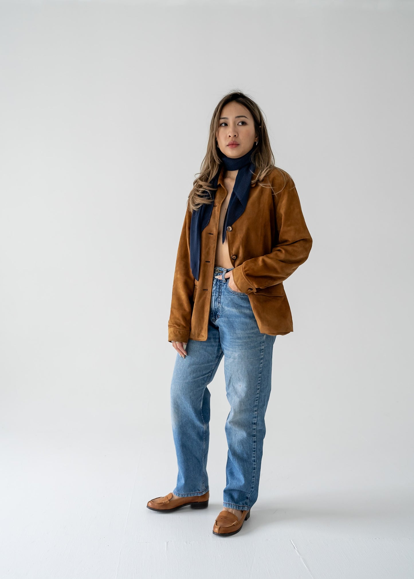 A person wearing a navy blue micro pleat crinkle scarf with a brown suede jacket and blue jeans.
