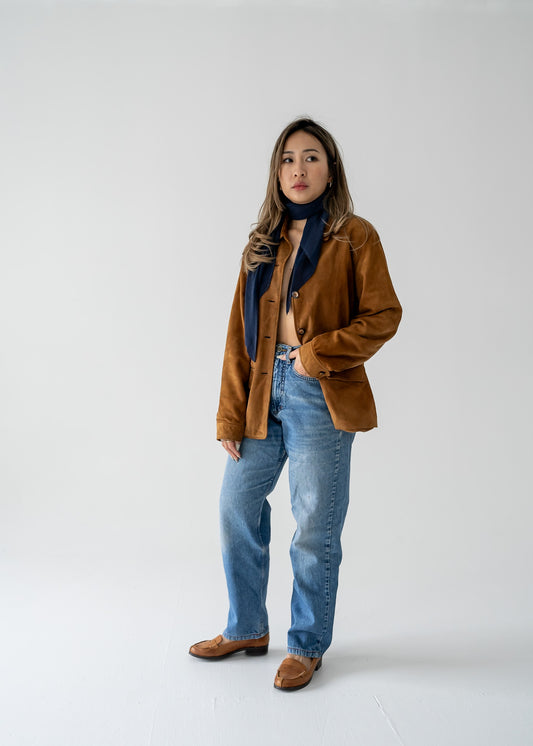 A person wearing a navy blue micro pleat crinkle scarf with a brown suede jacket and blue jeans.