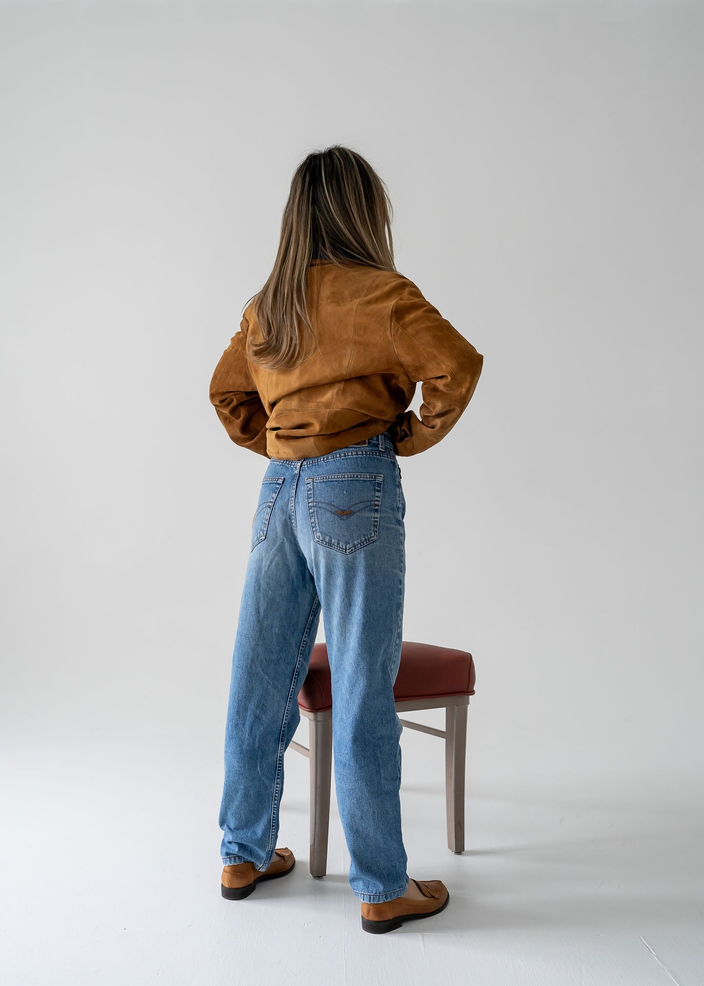 A person wearing a vintage tawny brown suede leather button-up collared jacket with blue jeans in front of a chair
