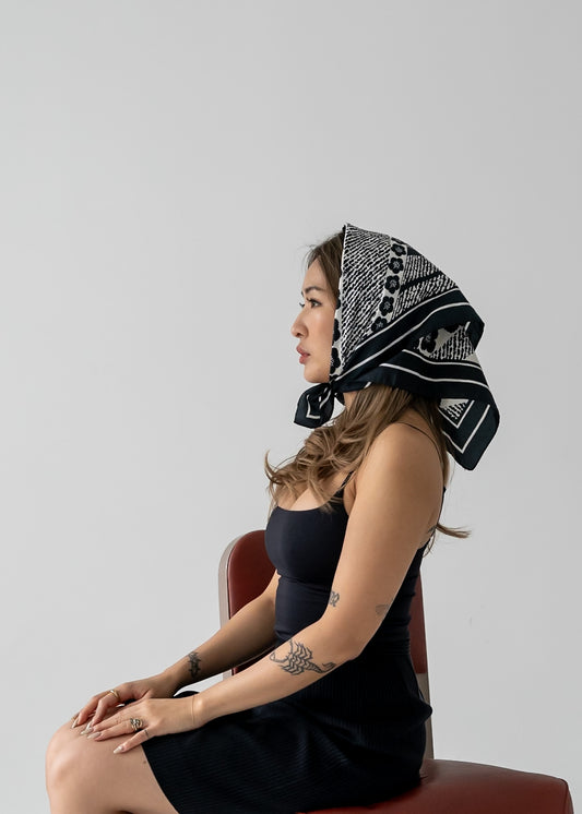 Woman sitting on a chair wearing a black and white floral patterned headscarf against a plain background