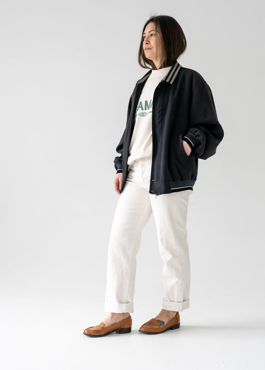 Person wearing a vintage lightweight navy bomber jacket, white shirt with text, and white pants on a light gray background