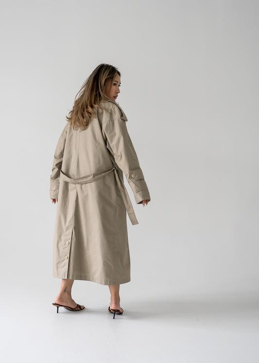 A person standing in a studio wearing a classic khaki long button-up trench coat with a double-breasted button closure, a classic collar, and a belted waist.