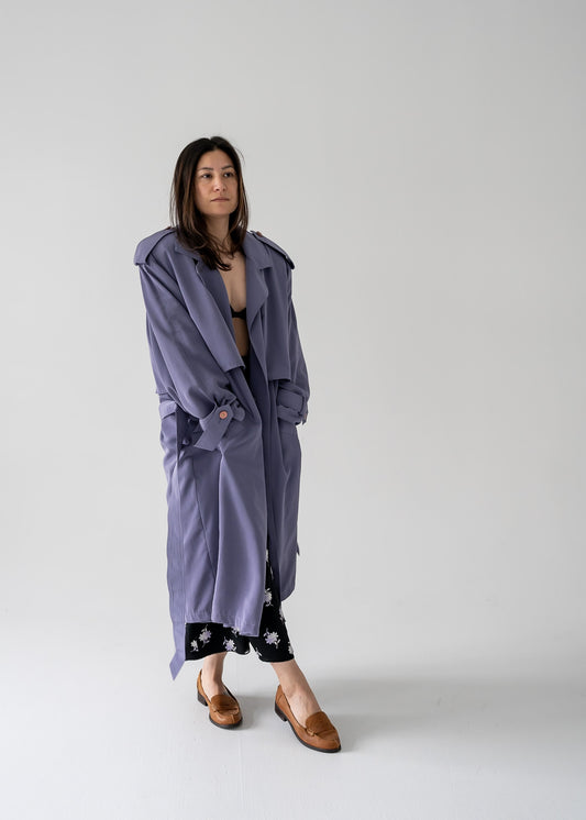 A person standing in a studio setting, wearing a heather purple long button-up trench coat with a notched lapel collar, sewn-in shoulder pads, slanted open pockets at hips, and a removable classic belt.