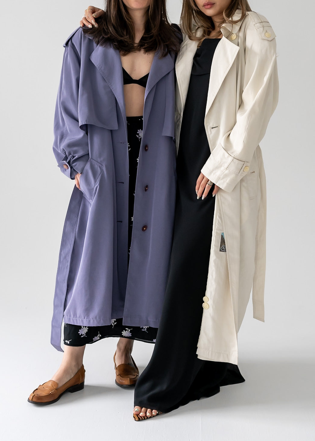 Two people wearing long vintage trench coats, one purple and one ivory, standing side by side.