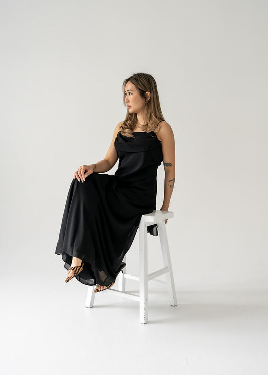 Woman in a floor length vintage black crepe dress sitting on a white stool against a plain background
