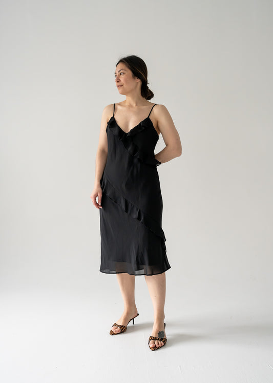 A woman standing wearing a vintage black spaghetti strap bias ruffle midi dress with a v-neck.