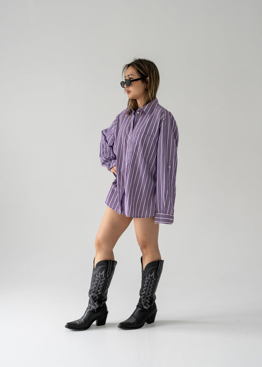 A person standing against a white background wearing a vintage purple and white striped long sleeve button-up shirt and black boots.