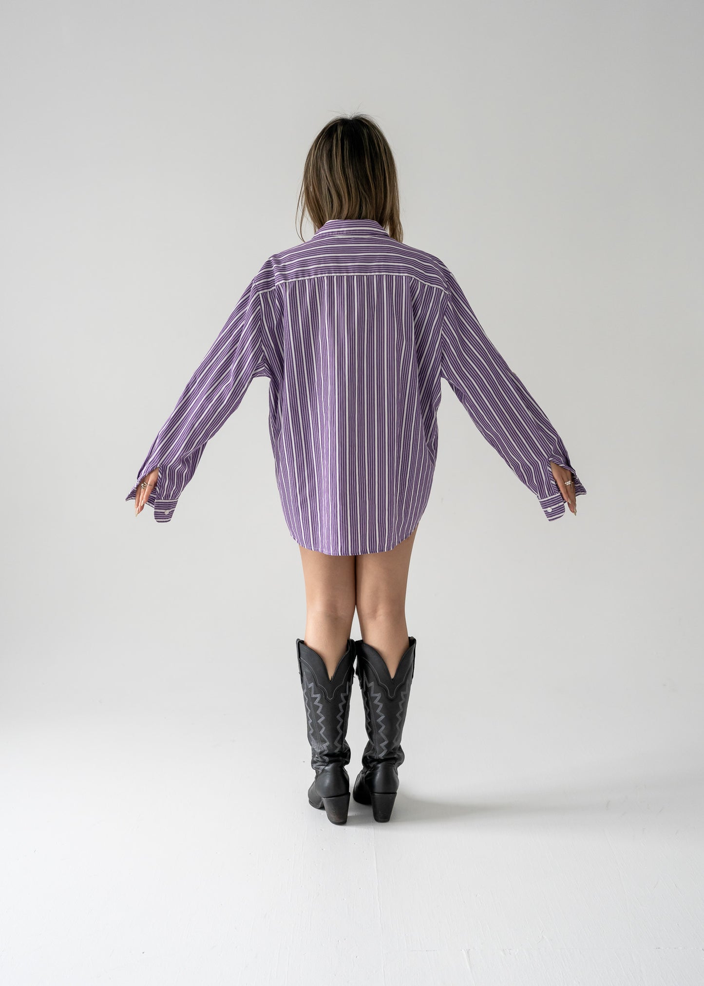 A person standing against a white background wearing a vintage purple and white striped long sleeve button-up shirt and black boots.