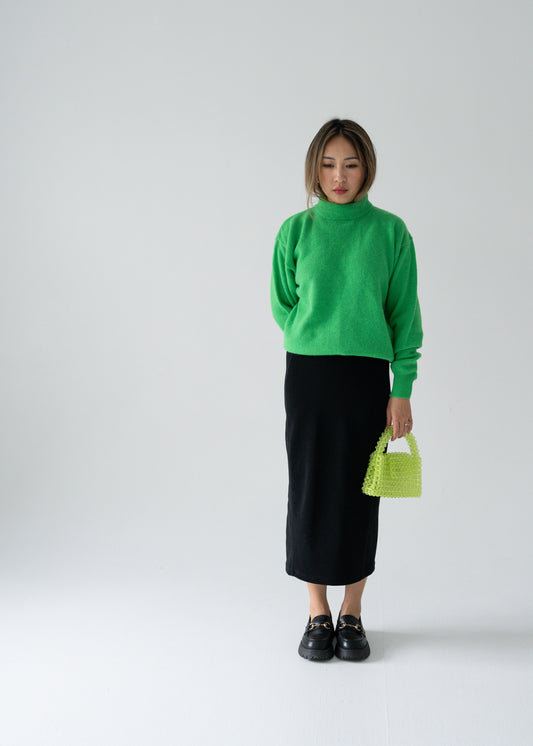 Person wearing a vintage candy green mock neck long sleeve wool Angora pullover sweater and black skirt holding a green bag against a white background