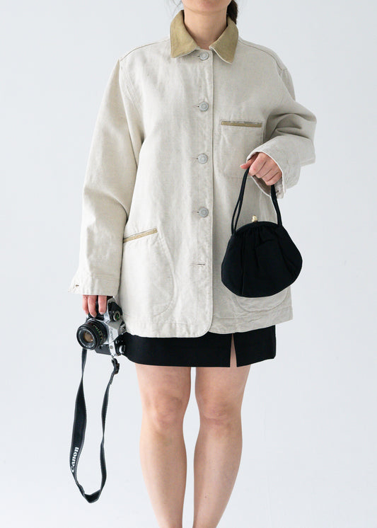 A person wearing a lightweight vintage flax linen and light moss green suede button up work jacket with a camera around the neck and a black skirt.