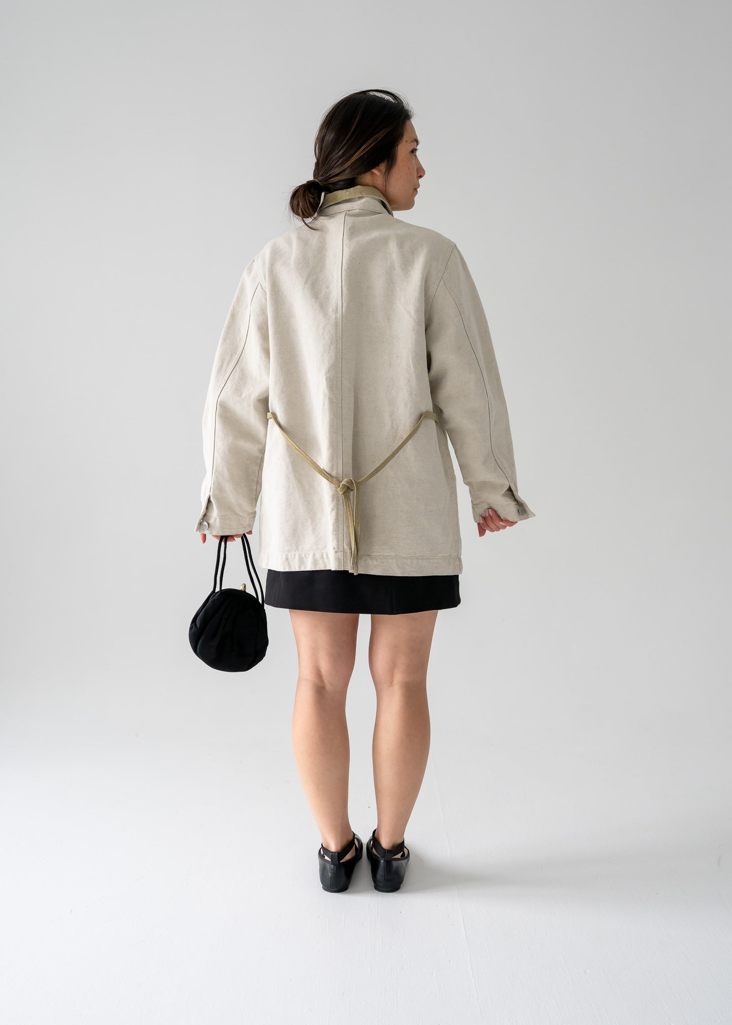 Person wearing a lightweight vintage flax linen and light moss green suede button up work jacket and black skirt holding a black bag on a plain background.