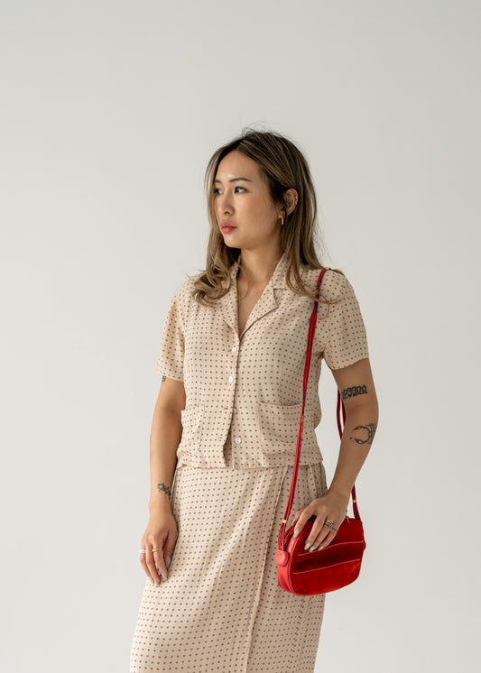 A person wearing a dotted dress holding a vintage strawberry red suede leather crossbody bag with small gold hardware.