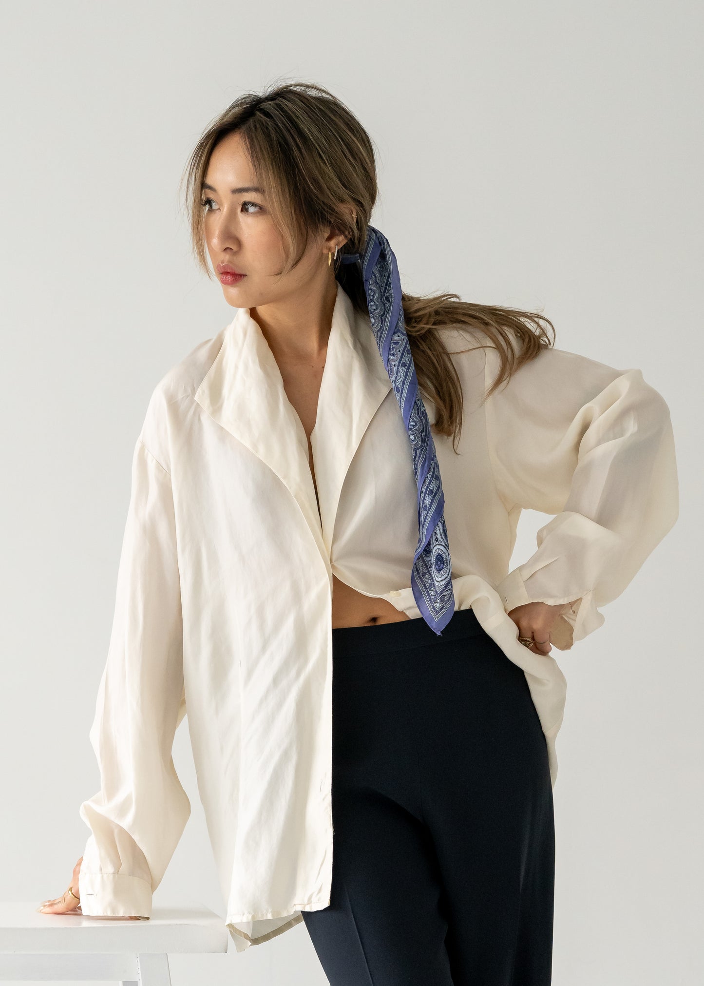 Woman wearing a white shirt and dark pants with a purple and blue silk scarf tied in her hair, standing against a plain background.