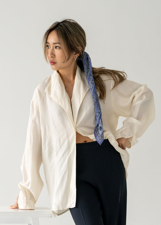 Woman wearing a white shirt and dark pants with a purple and blue silk scarf tied in her hair, standing against a plain background.