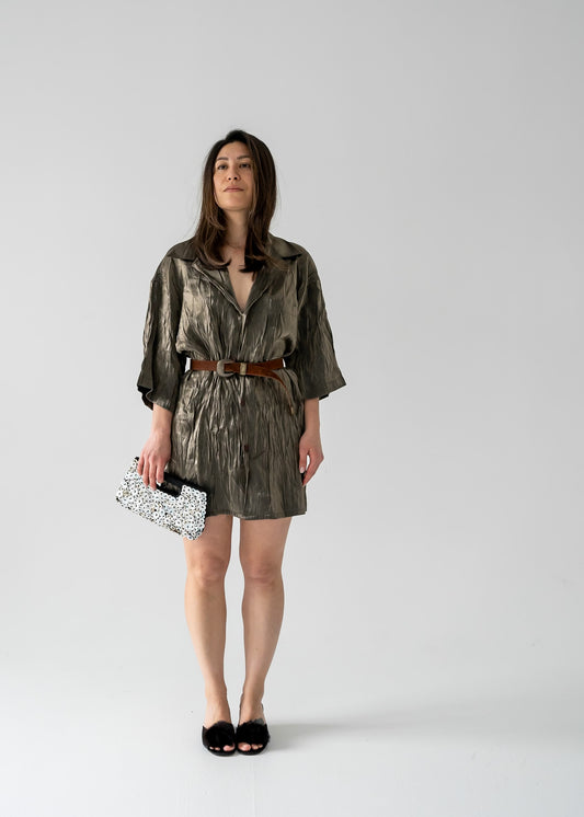 A woman standing in a side profile pose wearing a vintage green bronze full sheen textured lightweight short sleeve button up mini dress with a collared neck holding a vintage shell clutch.