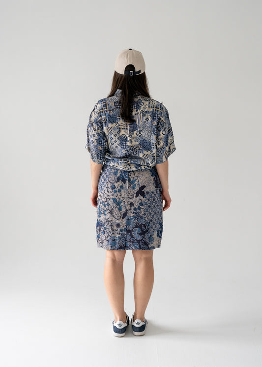A person standing against a white background, wearing a vintage multi-colored blue and cream abstract and floral pattern pure silk short sleeve button up blouse with a matching skirt