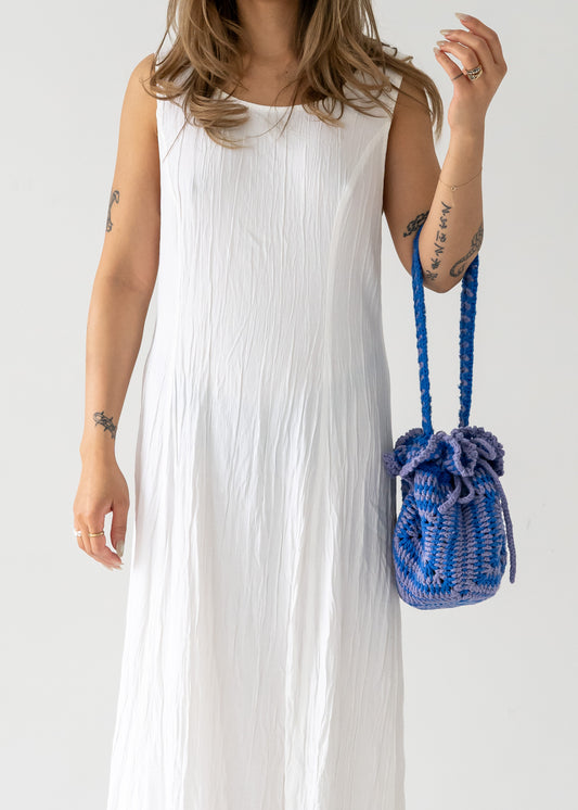 A person wearing a vintage sleeveless, ivory, crinkle texture floor-length dress with a princess seam, holding a blue woven bucket bag.