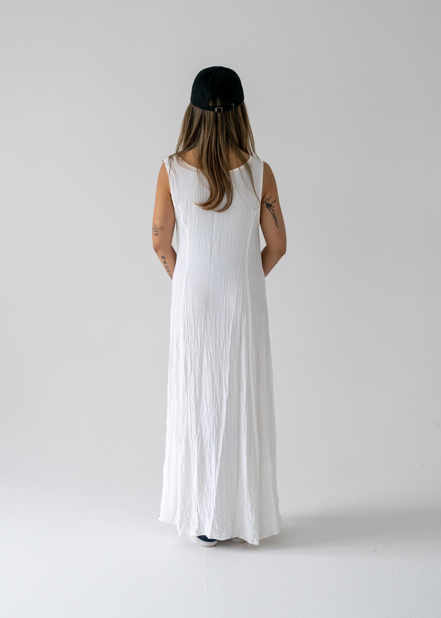 Person wearing a vintage ivory crinkle lightweight floor-length princess seam sleeveless dress and black cap on a plain background