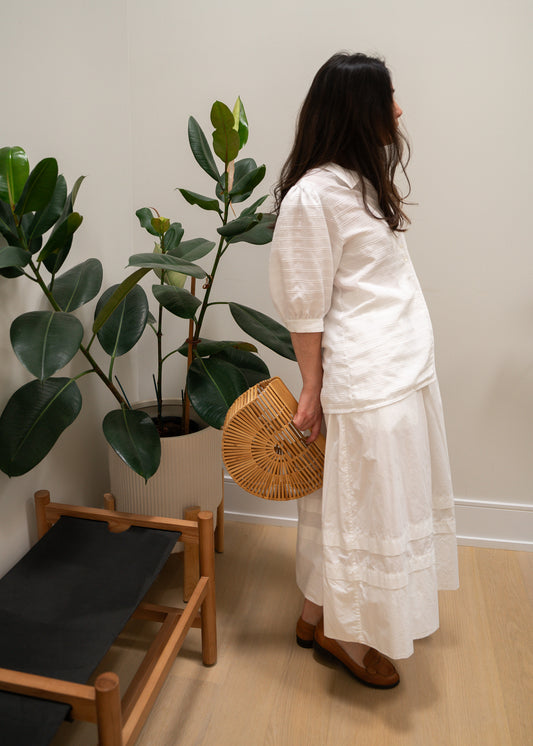 Woman in a white striped puffy sleeve blouse and white skirt holding a woven bag in a room with plants and a chair.