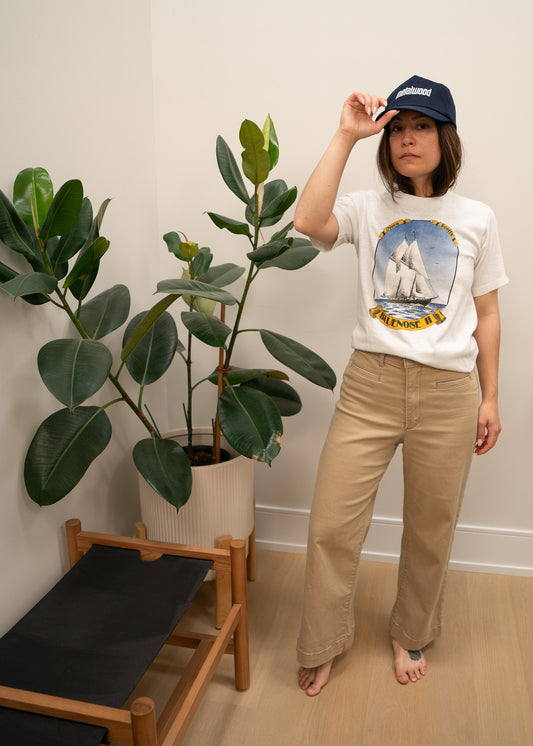 Person wearing a white t-shirt with a boat graphic design, blue hat and light pants, standing indoors with plants in the background.