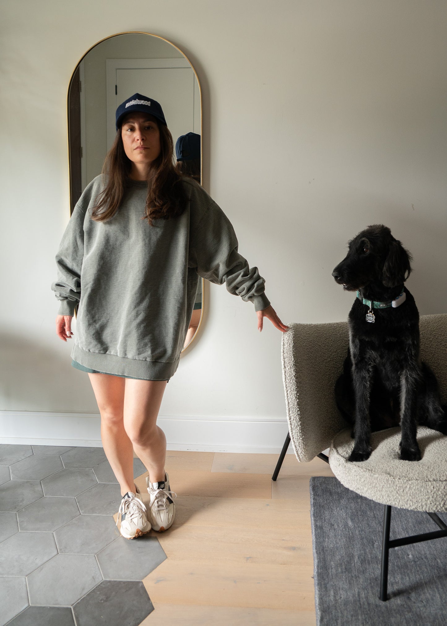 Woman in a grey green sweatshirt standing next to a black dog on a chair in a room with a mirror.
