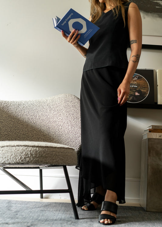 Person wearing a black sleeveless top and black skirt holding a book in a room with a chair and table.