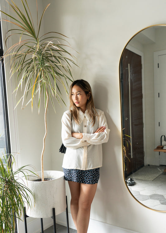 Woman wearing a white beaded blouse and navy floral skirt standing against a light wall, plant and mirror