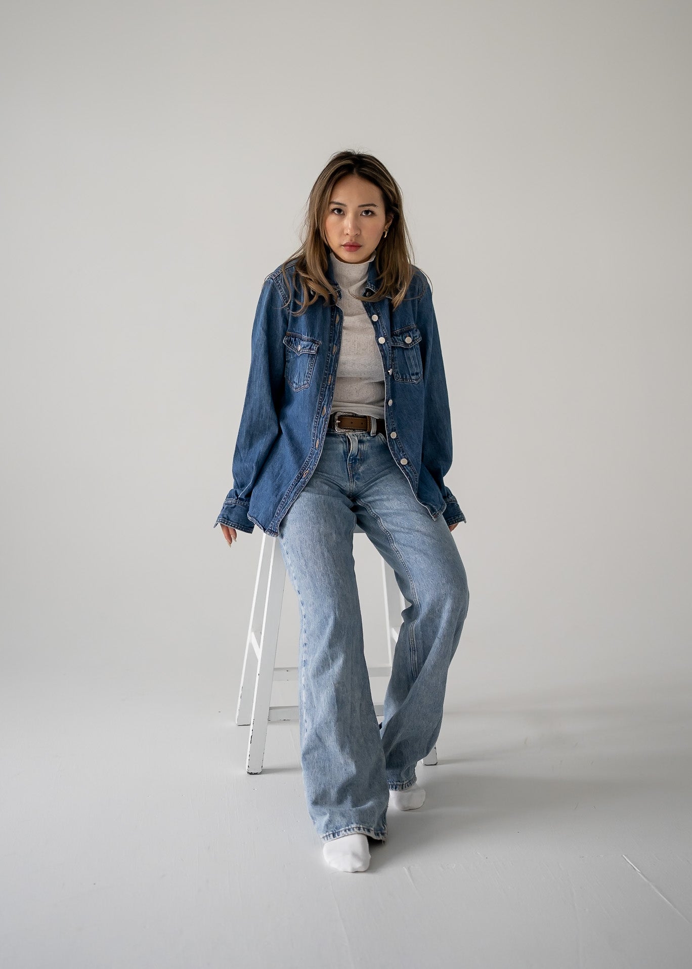 A person wearing a vintage medium wash denim button up long sleeve shirt with a round neckline, collar, and two buttoned flap pockets on the breast. They are also wearing light wash denim jeans and white shoes, standing against a white background.