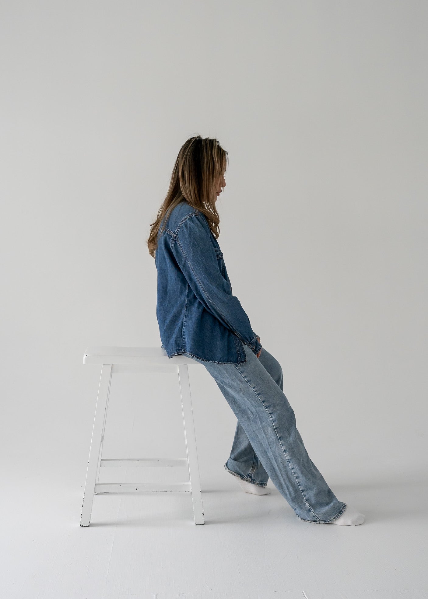 Person sitting on a stool wearing a vintage medium wash denim button up long sleeve shirt and jeans on a white background 