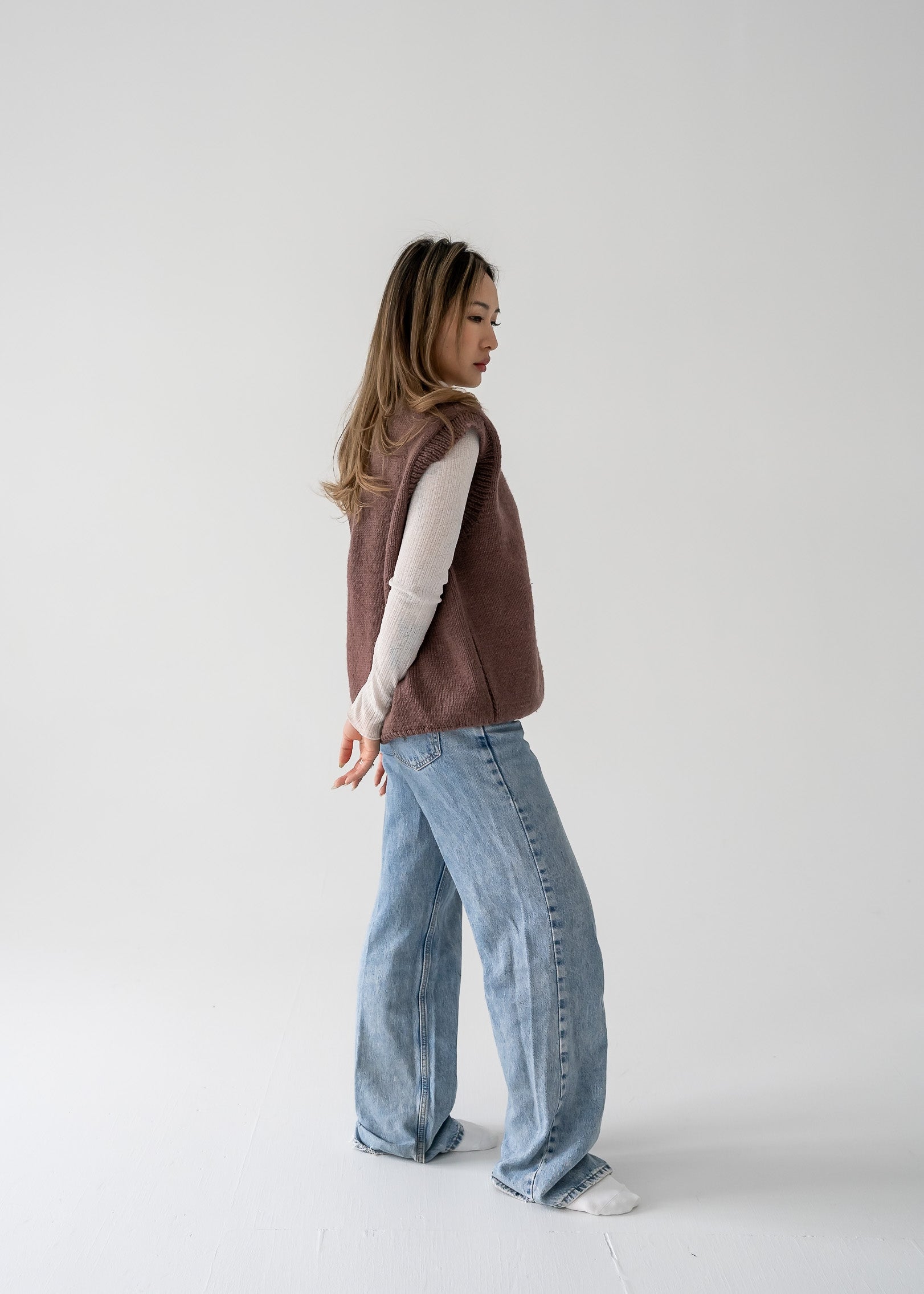 Person wearing a warm vintage brown thick knit V neck vest and blue jeans on a white background