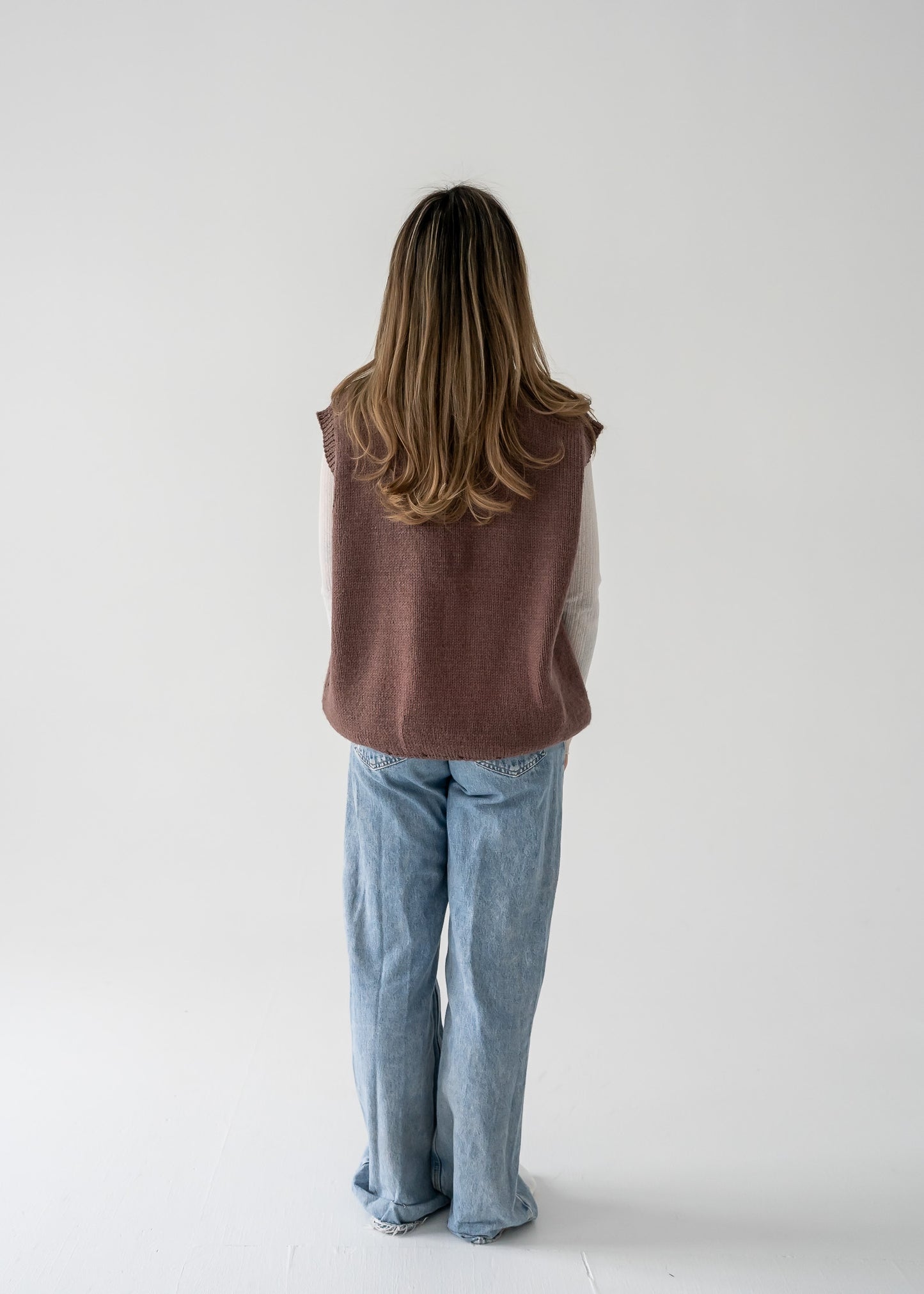 Person wearing a warm vintage brown thick knit V neck vest and blue jeans against a white background.
