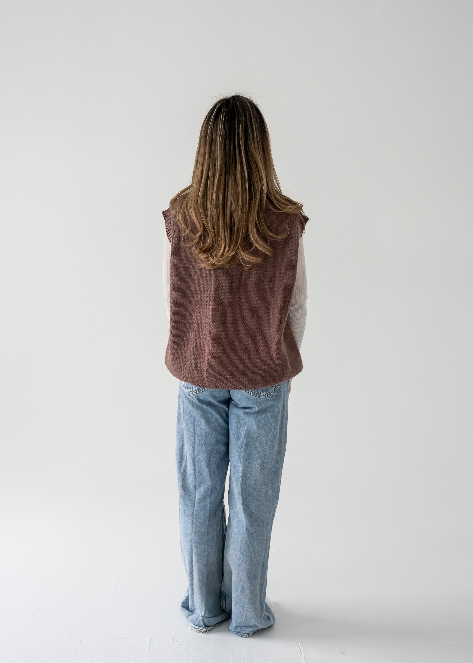 Person wearing a warm vintage brown thick knit V neck vest and blue jeans against a white background.