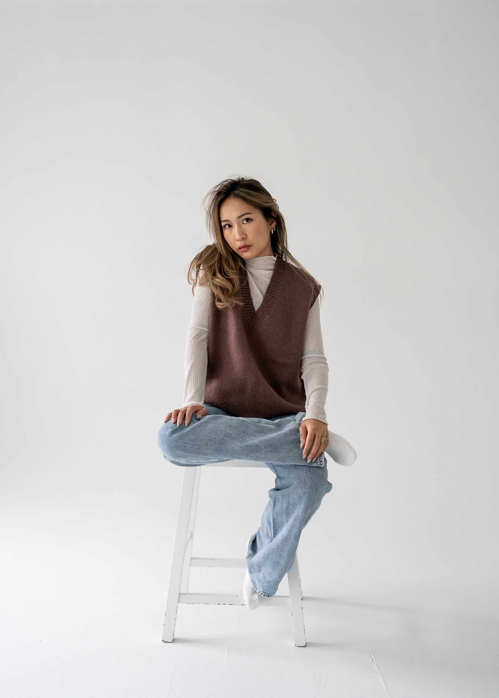 A person wearing a warm vintage brown thick knit V neck vest and light blue jeans, sitting on a white stool against a white background.
