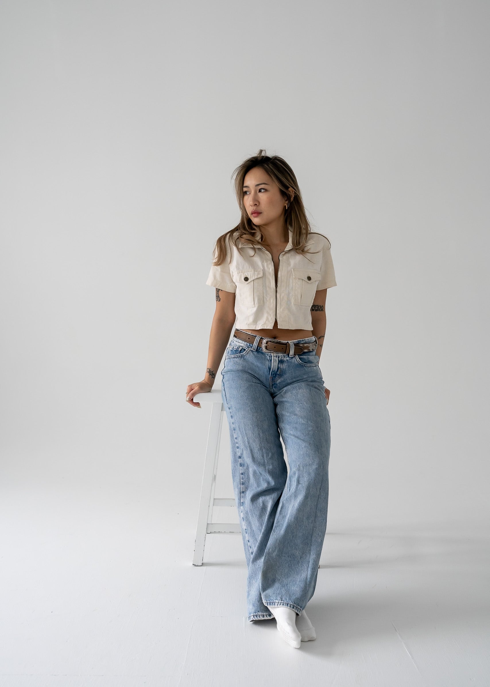 A woman standing wearing a vintage beige cropped linen zip-up collared short sleeve jacket style top with blue jeans and white shoes.