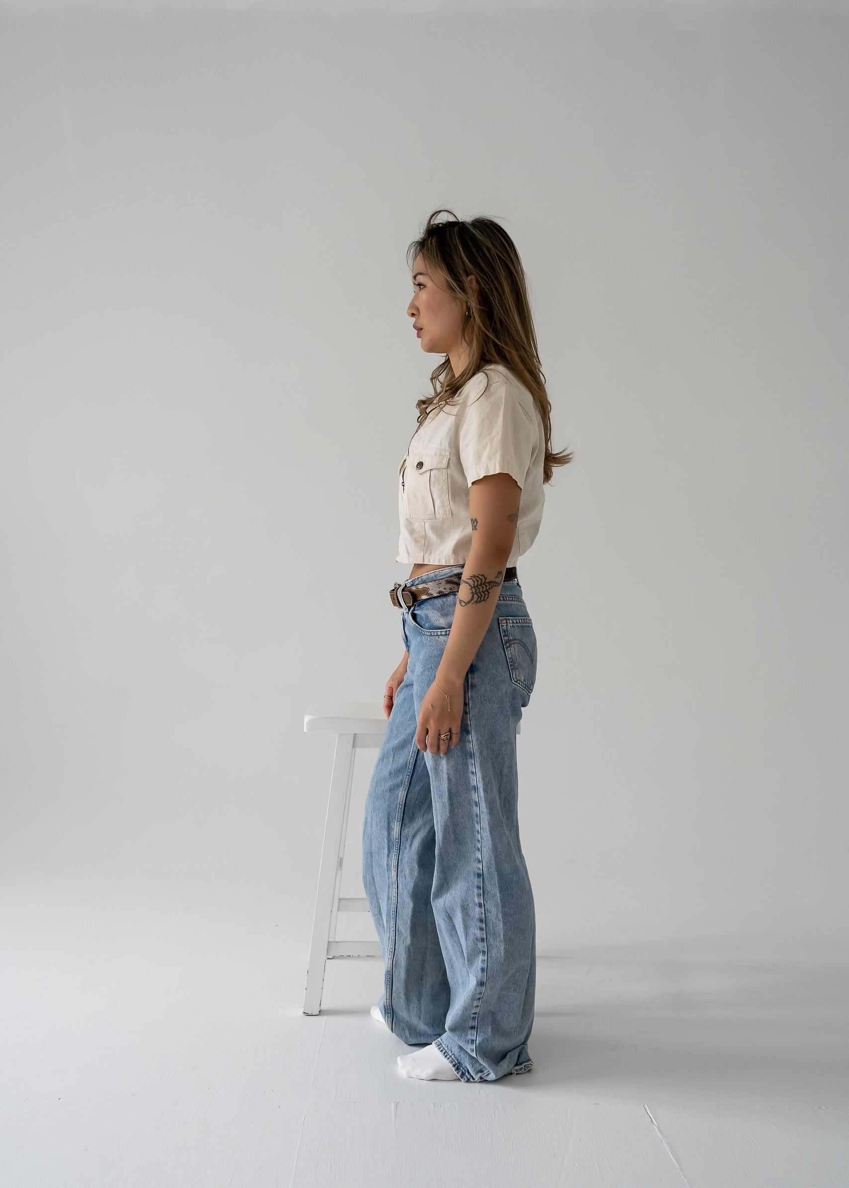 Person wearing vintage beige cropped linen zip-up collared short sleeve jacket style top and blue jeans standing in a studio.