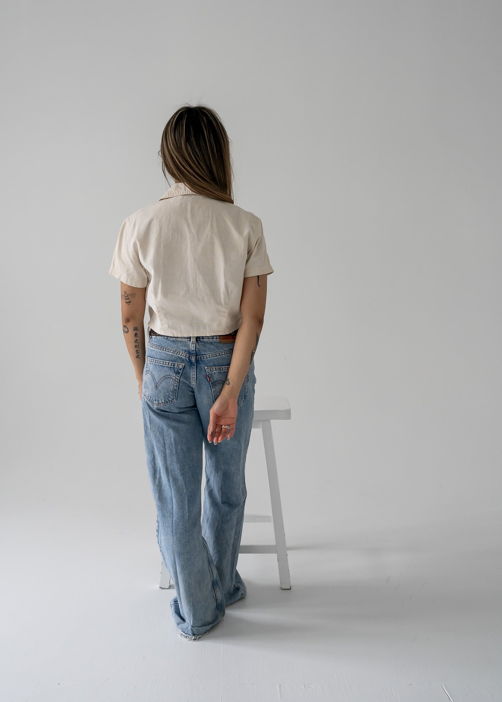 Person wearing a vintage beige cropped linen zip-up collared short sleeve jacket style top and blue jeans standing in a minimalistic setting.