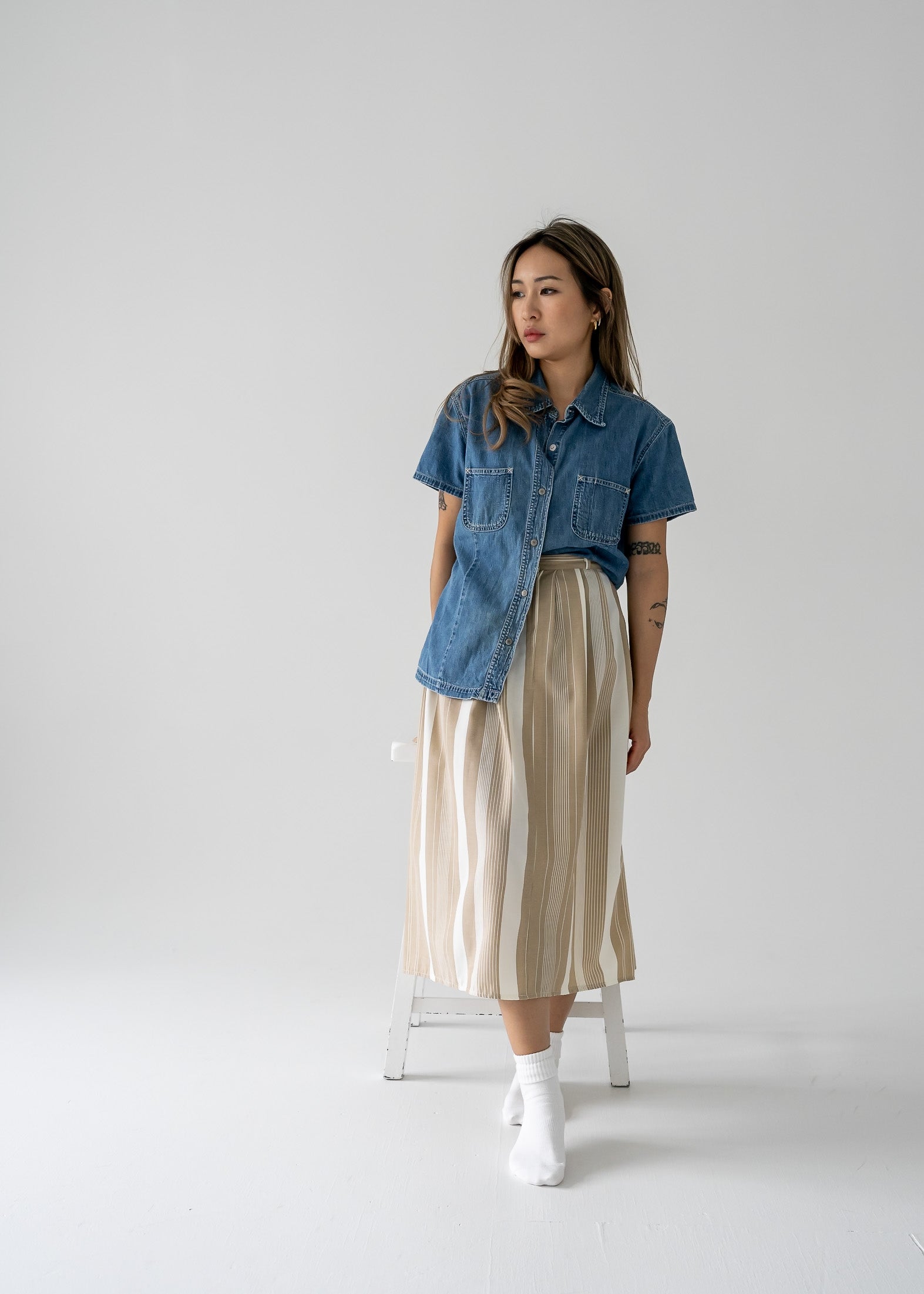 A person wearing a vintage medium wash denim short sleeve shirt with metal snap buttons and a beige skirt.
