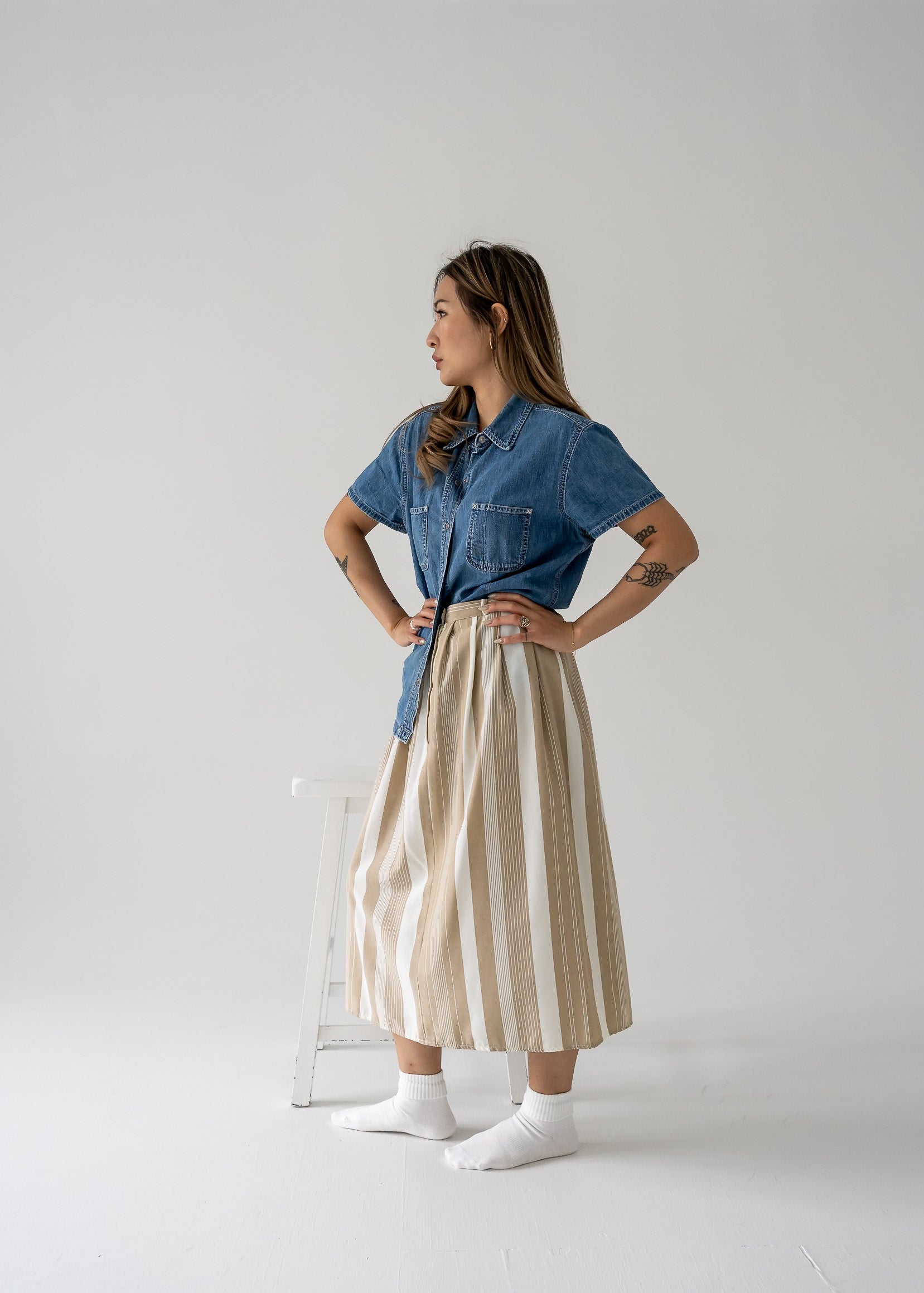 A model wearing a beige and cream vertical stripe pleated waist midi skirt with a denim shirt standing against a white background.