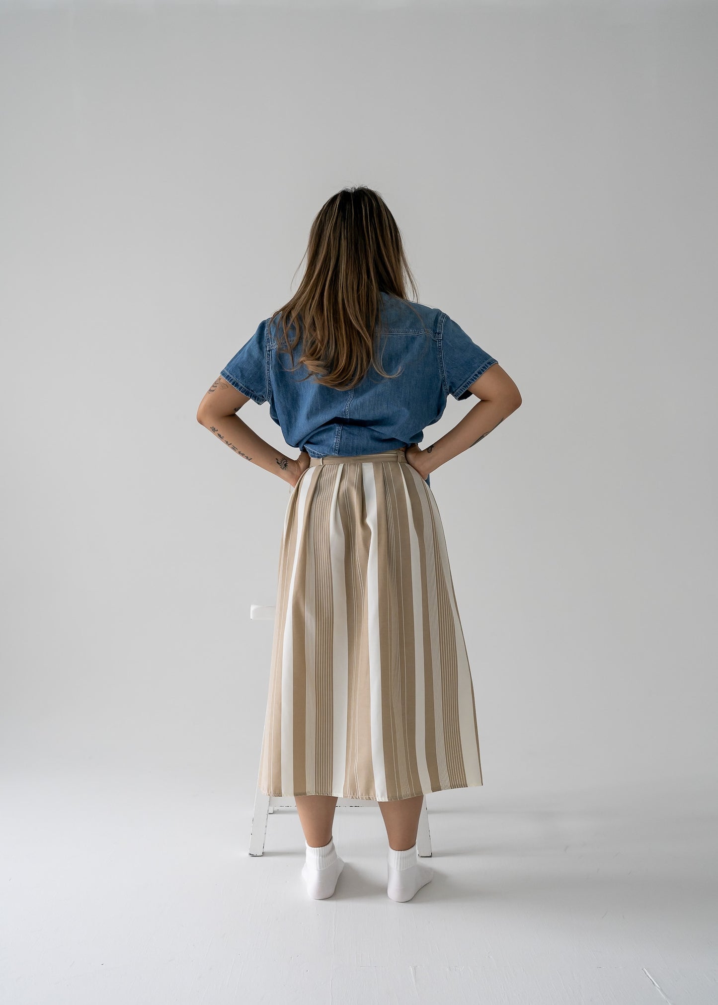 A model wearing a beige and cream vertical stripe pleated waist midi skirt with a denim shirt standing against a white background.