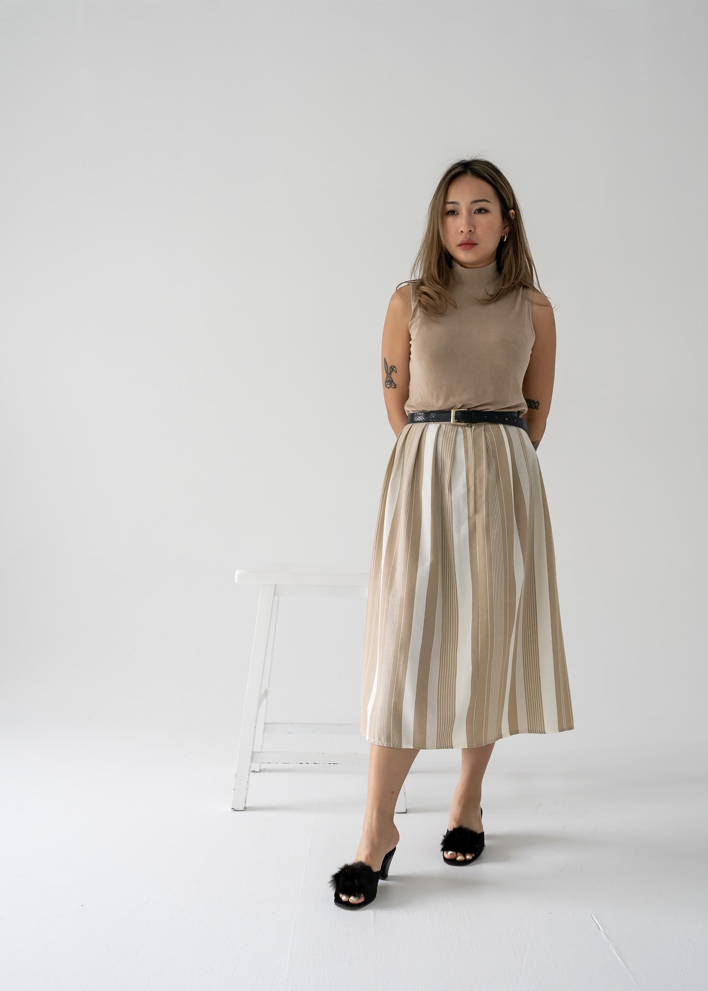 Woman in beige sleeveless top and beige and white striped pleated skirt standing against a white background