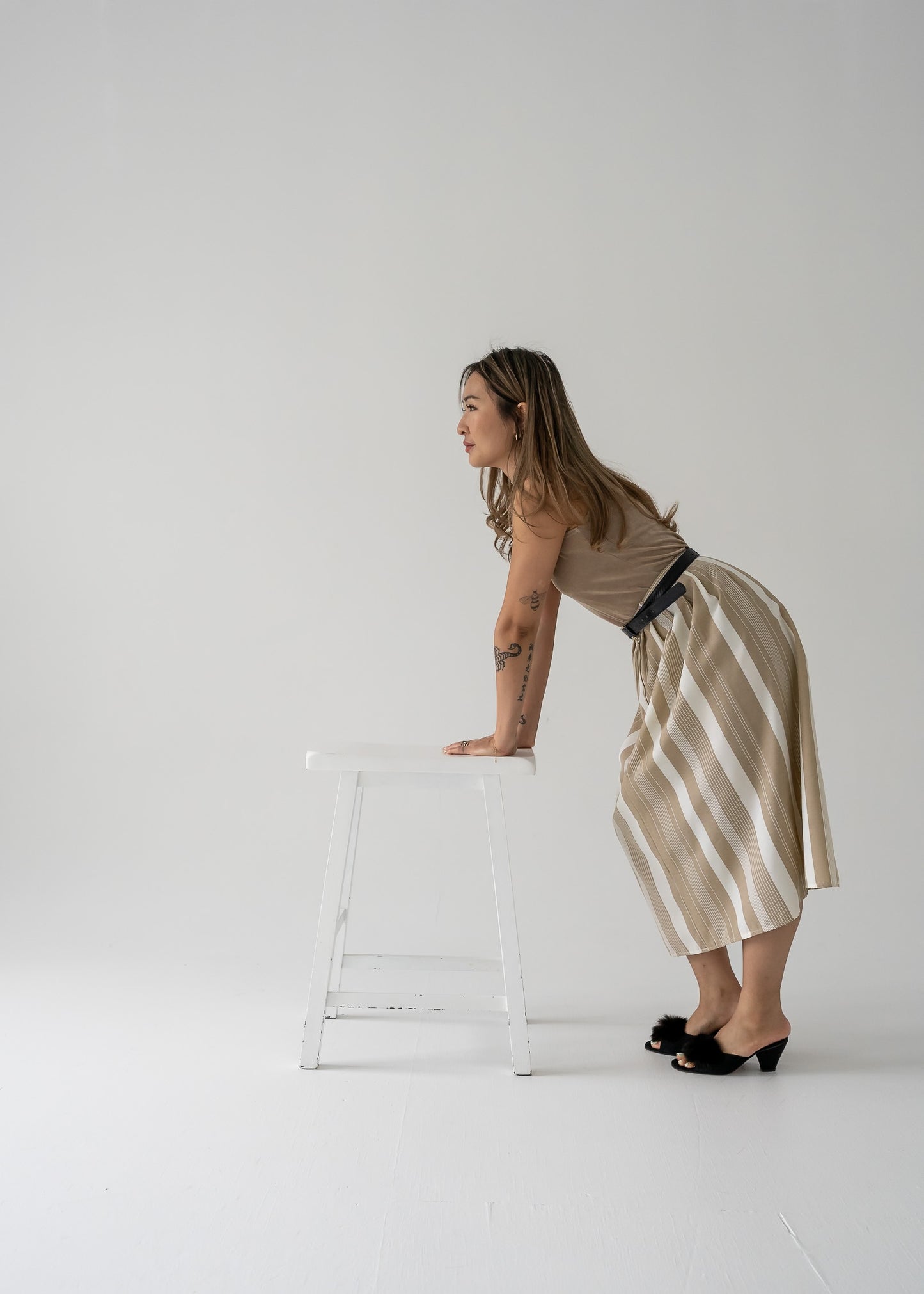 Woman in beige sleeveless top and beige and white striped pleated skirt leaning against a stool against a white background