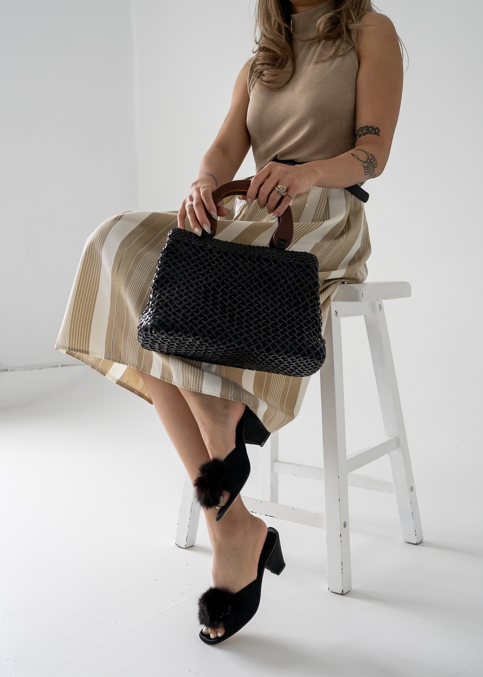 Woman sitting on a stool holding a black handbag, wearing a beige tank top and striped skirt and black pom pom kitten heel shoes.