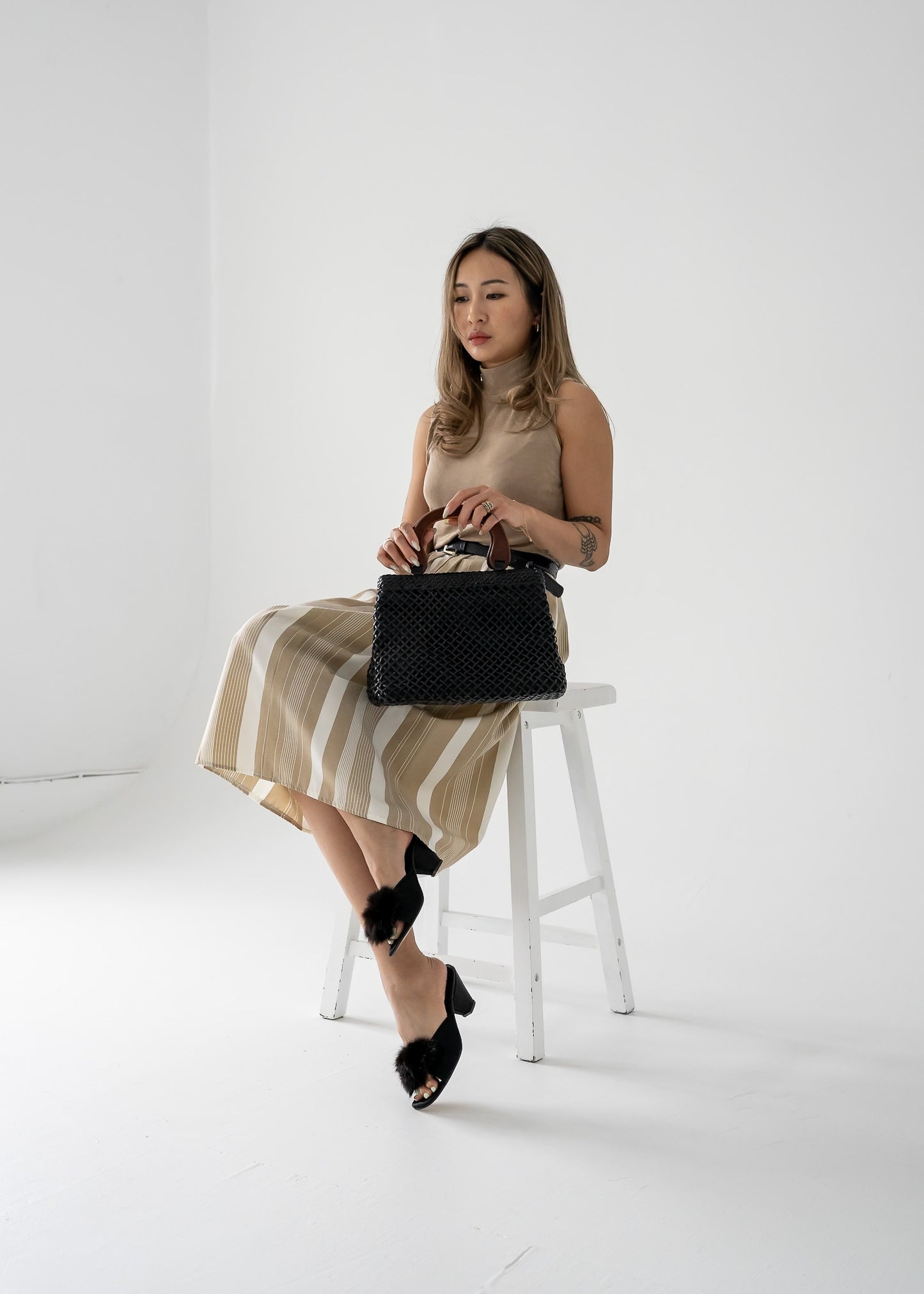 Woman wearing a vintage beige tank top and striped beige skirt sitting on a stool holding a black handbag in a minimalistic setting