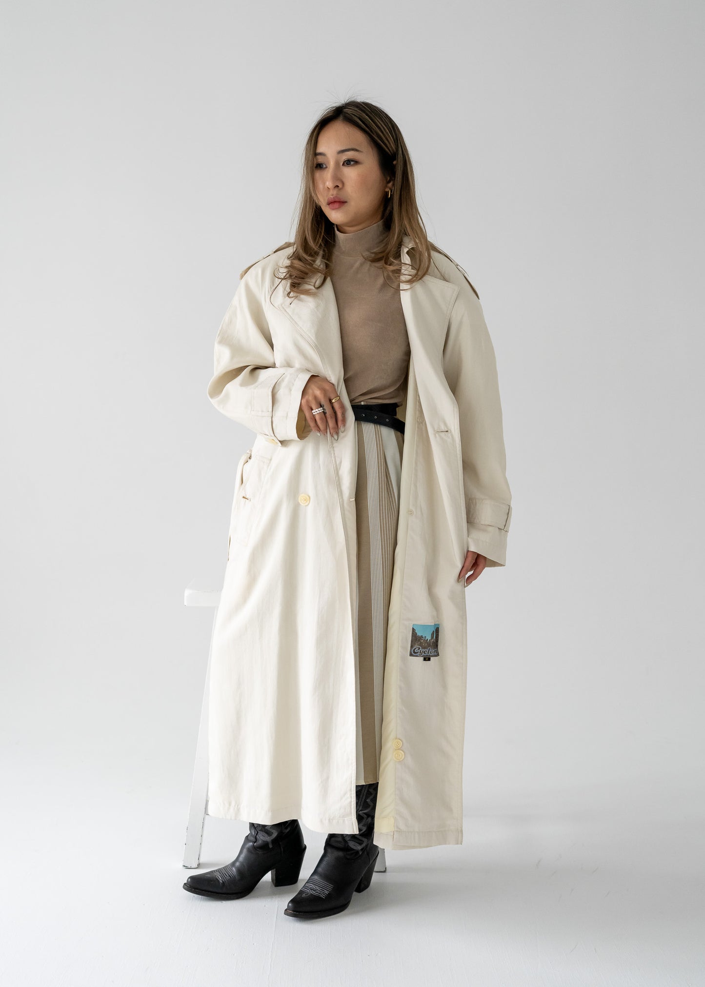 A female model wearing a vintage cream-colored, double-breasted, belted, full-length trench coat with a rounded collar and button-up front.