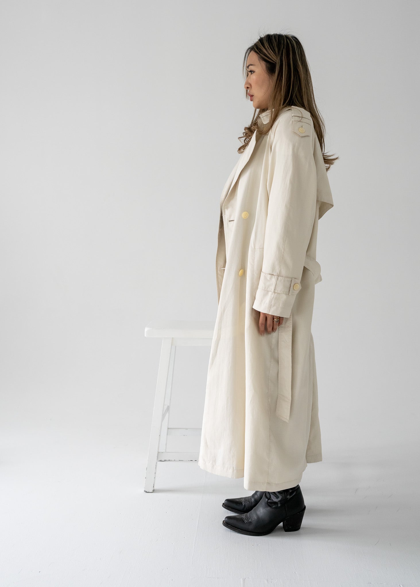 Woman wearing a vintage cream-colored, double-breasted, belted, full-length trench coat  in a minimal white studio.