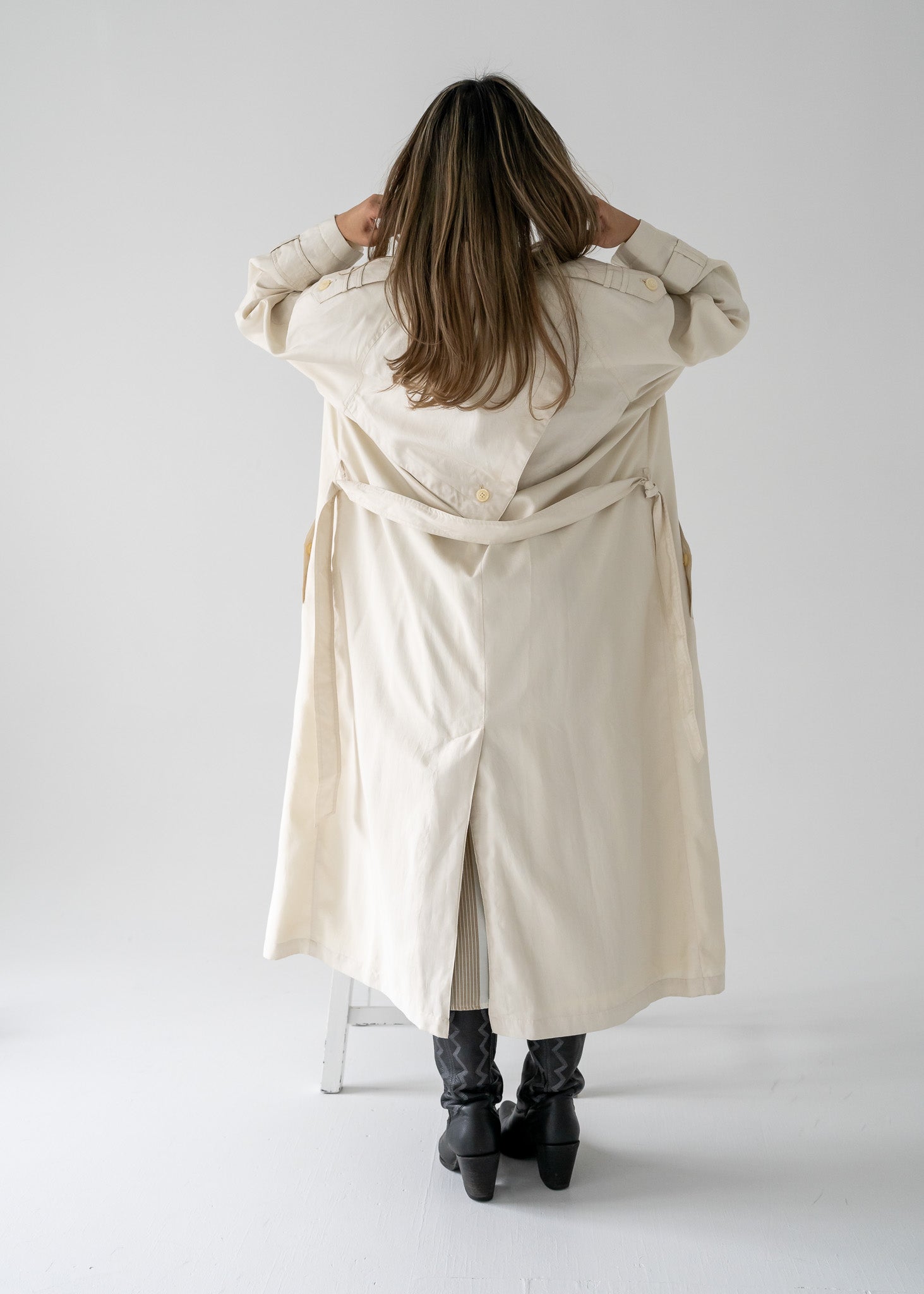 Person wearing a vintage cream-colored, double-breasted, belted, full-length trench coat on a white background